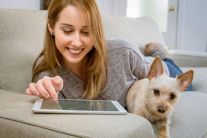 Girl looking at her tablet on a couch smiling with her dog beside her.