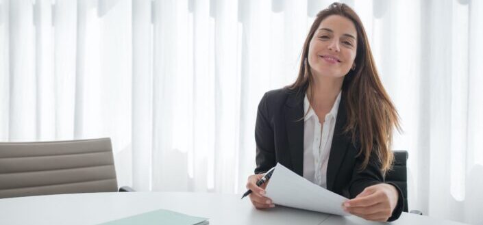 woman smiling in black blazer holding white paper and pen 