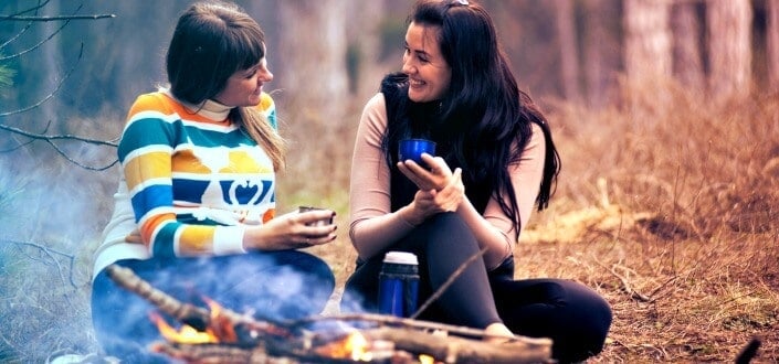 Two ladies enjoying coffee while sitting on a dry grass along the woods