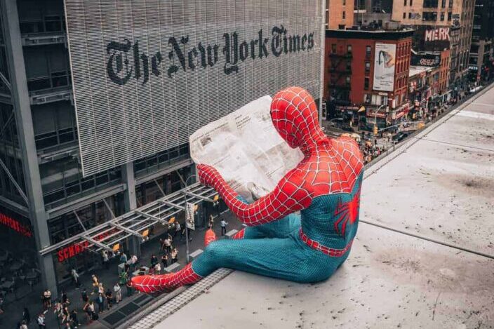 A guy dressed up in Spiderman costume sitting on a building rooftop while reading newspaper
