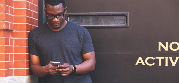 Man Leaning on Brown Brick Wall Holding Smartphone