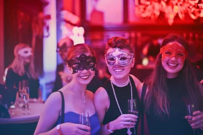 ladies in a masquerade party posing for a picture while holding their glass of wine - Halloween trivia