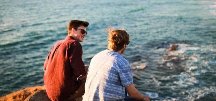 Two guy friends sitting on a beach rock, talking.