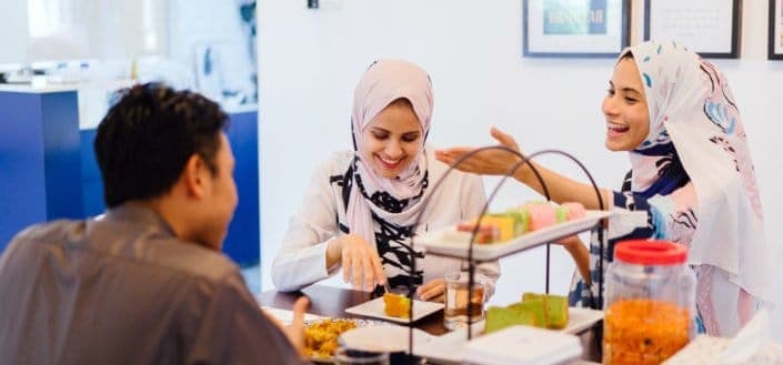 Two women in hijab, talking to a guy while eating.