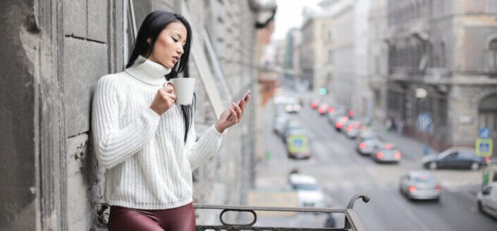 Girl reading text on her phone on a ledge with a cup of coffee.