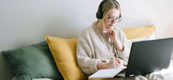 Girl writing on a notebook while listening to music on headphones and laptop in front of her
