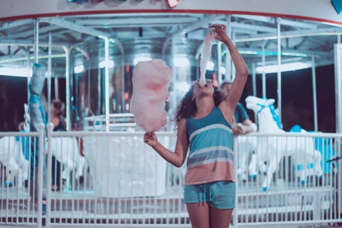 A person eating a large cotton candy in front of a carousel