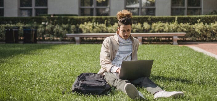 Guy sitting on grass with laptop on his lap and bag beside him.