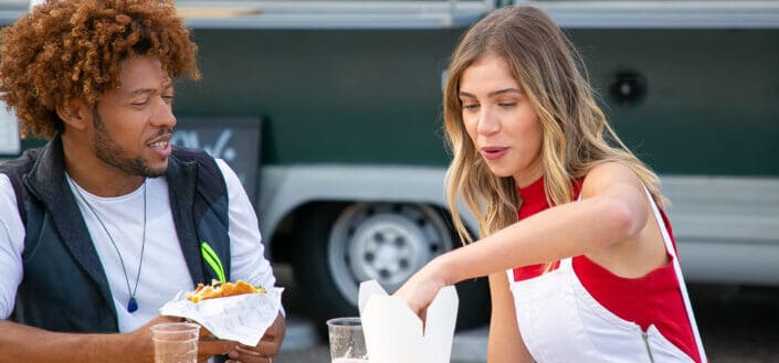 Hungry young multiracial couple drinking beer and eating fast food near van cafe