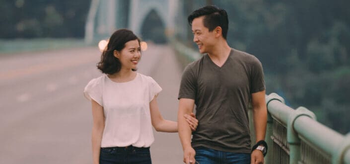 Cheerful young ethnic couple with dog strolling on bridge near green mountains