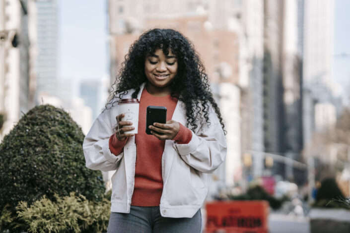 smiling woman with coffee browsing smartphone on street