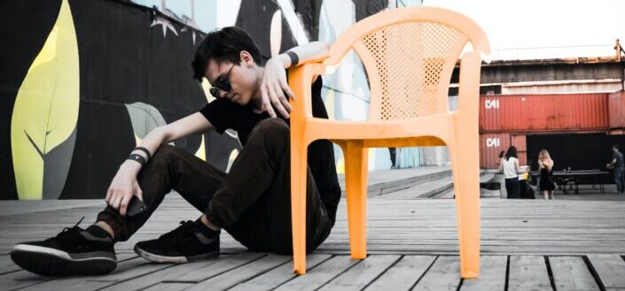 man sitting on floor beside empty orange plastic armchair at daytime