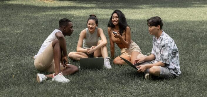 A group of friends with their gadgets sitting on a grass while having some fun conversation