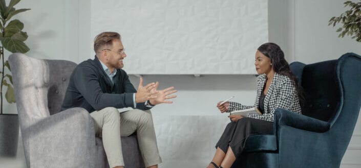 A man being interviewed by a woman while they both sitting on an office couch