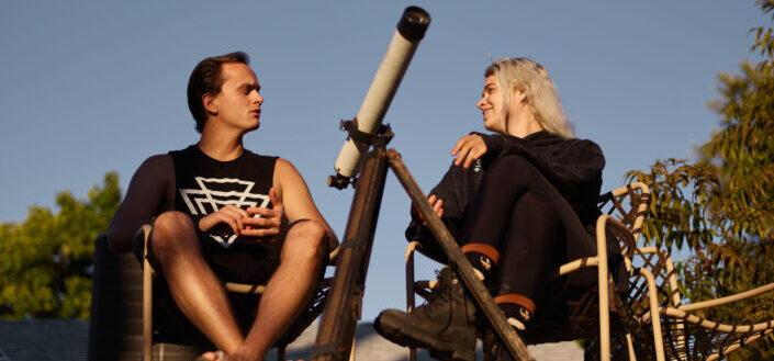 A man and a woman in black clothes sitting outdoors near a telescope