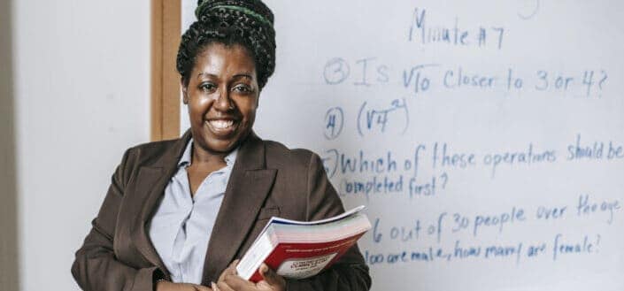 Cheerful black female teacher with workbooks standing near whiteboard