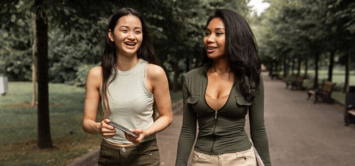 Positive young diverse women walking along path in park with gadgets in hands