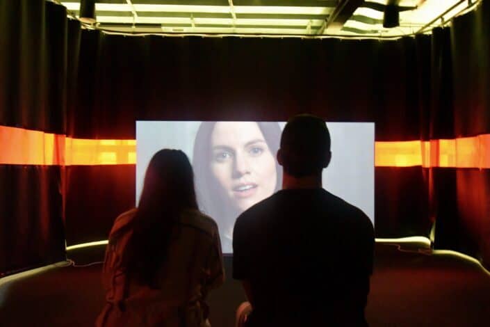 Two people mesmerized by a woman on a screen