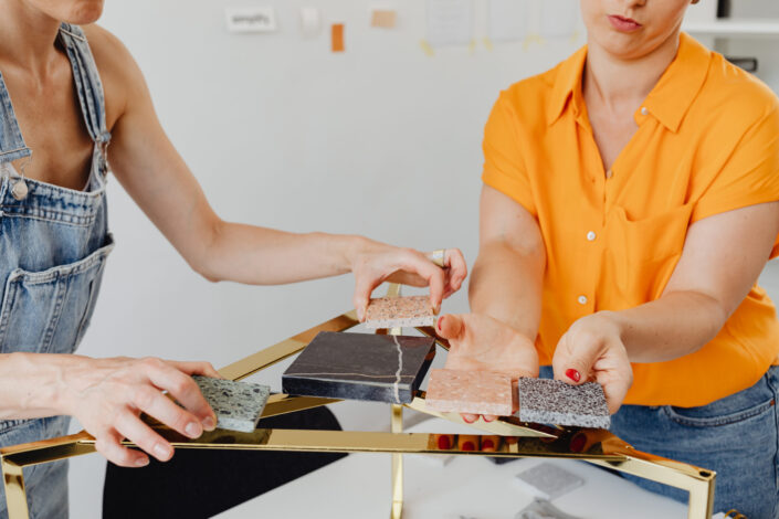 Woman in orange shirt and woman in denim overall holding pieces of marble beside golden table stand