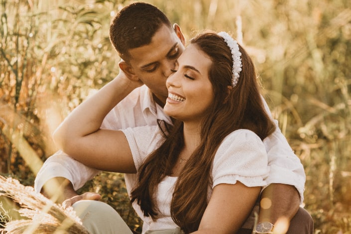 Ethnic boyfriend kissing cheerful girl while sitting together in grass