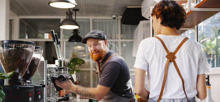 Smiling barista speaking with unrecognizable partner against coffee machine