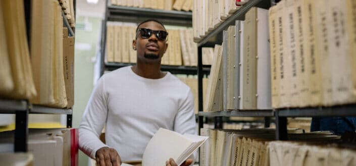 Man in white sweater wearing sunglasses while holding a book in library