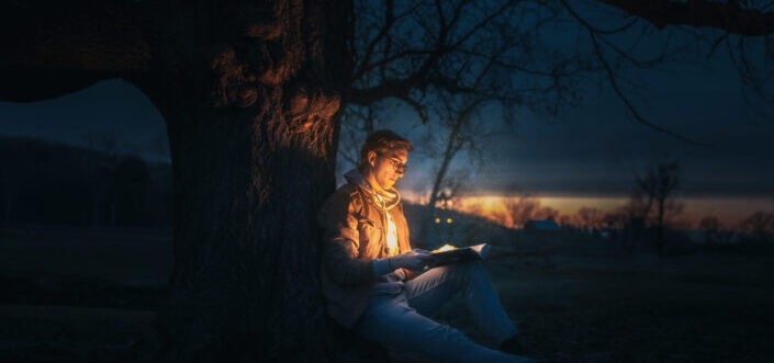 Man sitting under a tree reading a book during night time