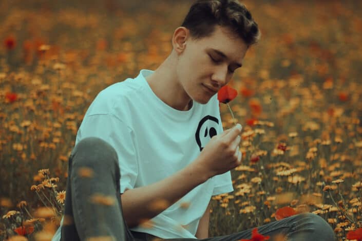 Teenage boy sitting on a flower field and smelling a poppy