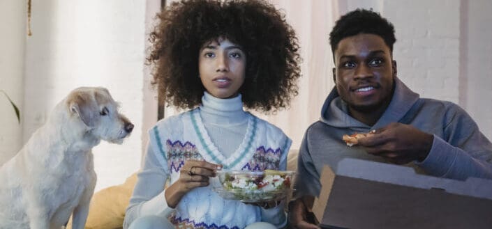 Focused young black couple having lunch and watching tv on couch near curious dog