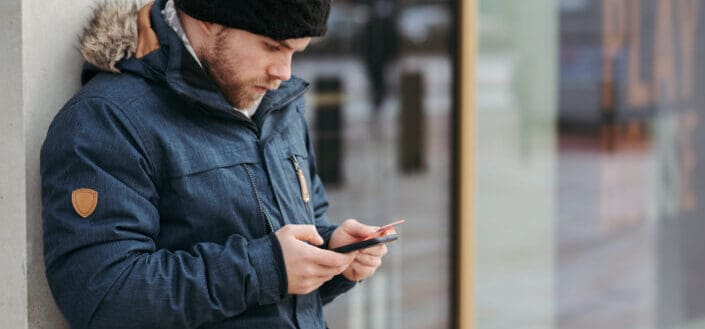 Serious man leaning on wall of building and using smartphone on street