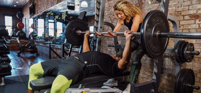 Lady gym trainor assisting a guy lifting a barbell