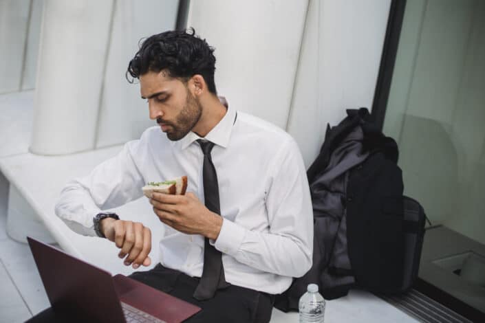 Man in a Corporate Attire Looking at His Watch While Eating a Sandwich