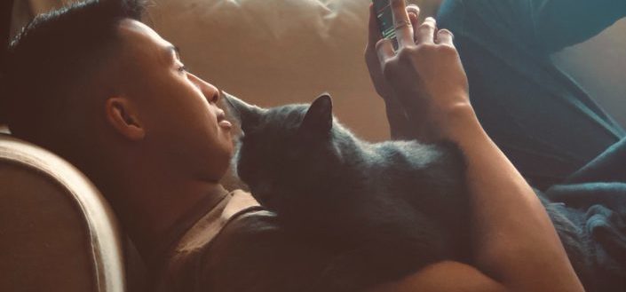 Guy on a couch with his cat, browsing his phone.