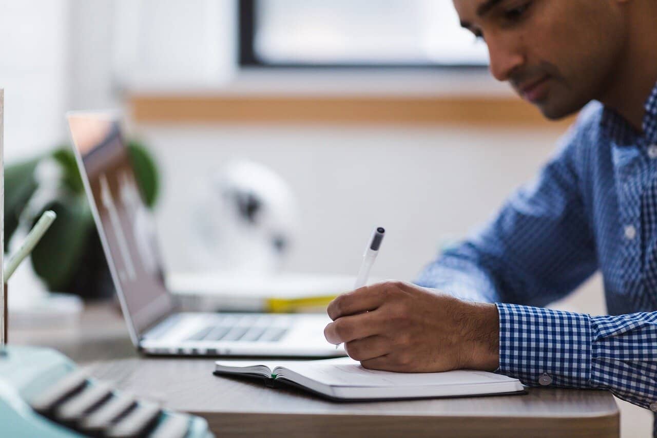 man writing on notebook while using laptop