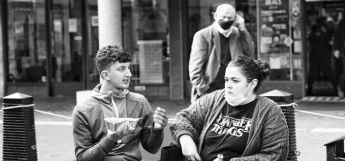 A young boy is having lunch and on a conversation with a young lady wearing a stranger things t-shirt with a middle-aged man wearing a black face mask because of COVID-19 is passing by at the background.